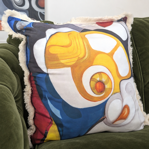 A square throw pillow with a colorful abstract face print, resting on an olive green velvet sofa. The pillow features a cream-colored fringed edge and a bold graphic design with yellow, blue, white, red, and gray shapes.